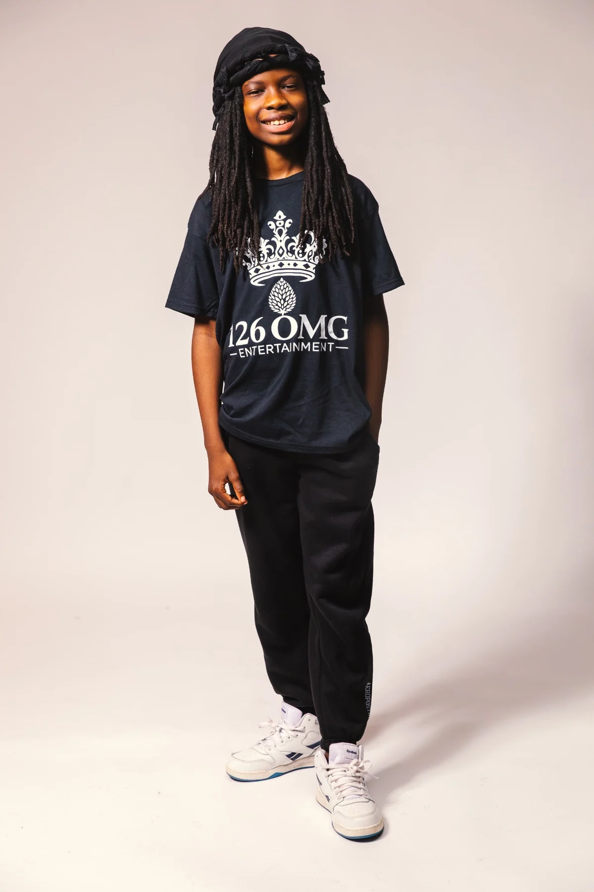 A son in a 126 OMG Entertainment shirt with long locs and white sneakers